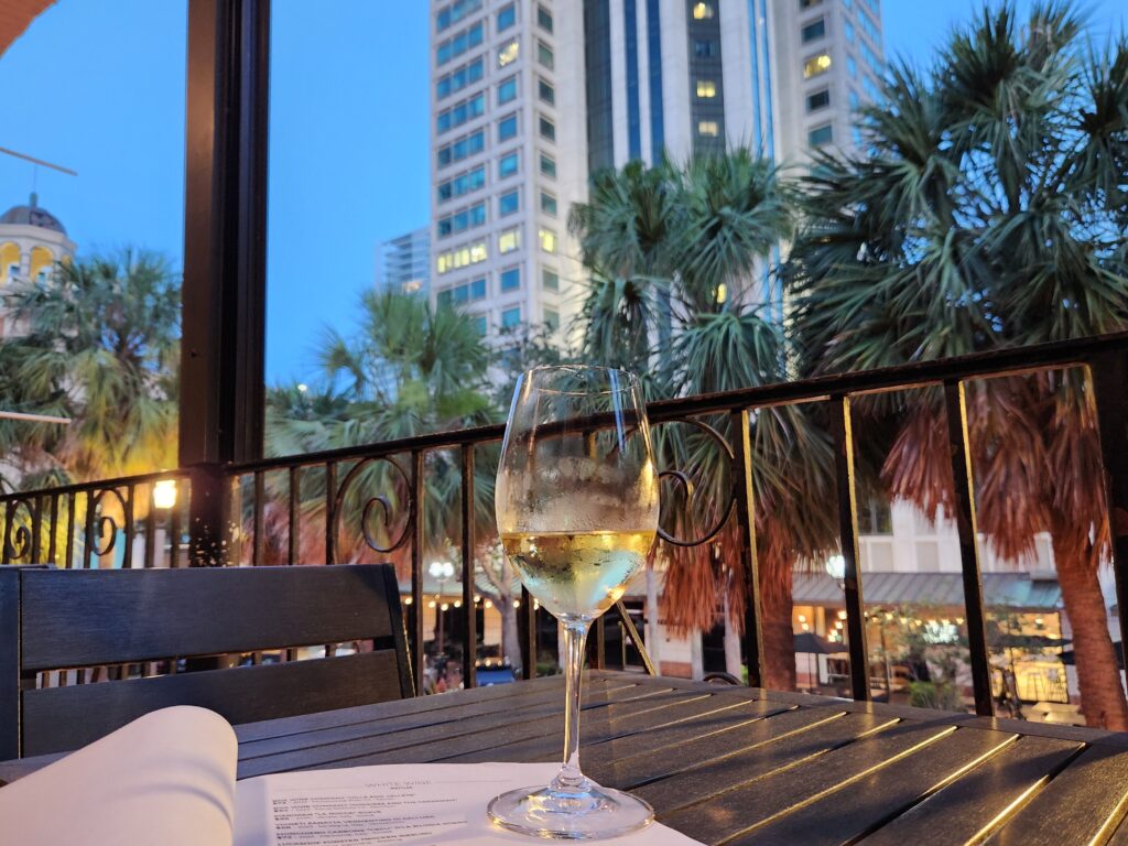 A glass of wine rests on a balcony table in St. Pete, with palm trees and a condo building rising in the background.