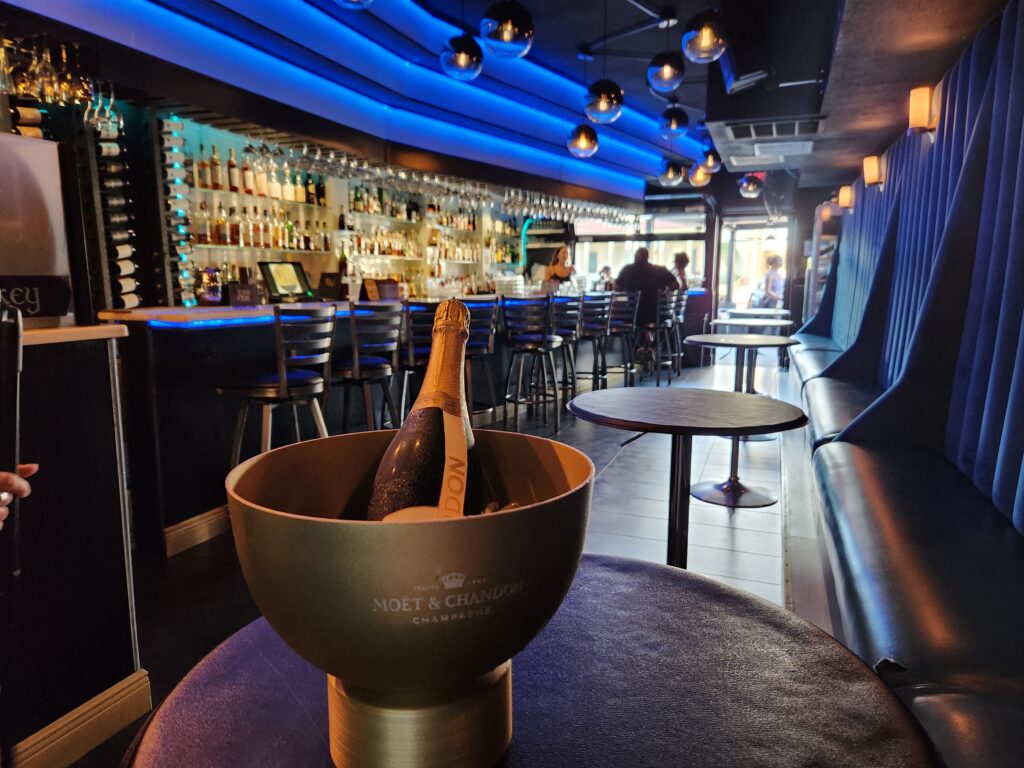 Moët & Chandon Champagne in a gold ice bucket on a table inside Flûte & Dram, with blue lighting and bar seating in the background.